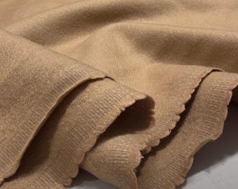 May include: Close-up of a tan fabric with a scalloped edge. The fabric is folded, showing the texture and color variations. The material appears soft and possibly knitted or woven, suitable for apparel or home decor.