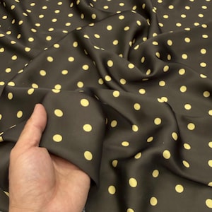 Op de afbeelding: Een donkerbruine stof met een herhalend patroon van kleine, lichtgele stippen. Het materiaal heeft een glad, licht reflecterend oppervlak met zachte plooien en draperingen die zorgen voor een gevoel van diepte en textuur in de stof.