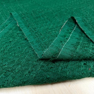 May include: Close-up of a textured, emerald green fabric. The fabric has a woven appearance with visible threads and a slightly uneven surface. The edges of the fabric are visible, showing a raw, unfinished edge with white stitching.