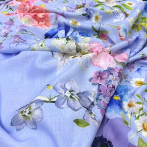 Peut inclure: Un tissu bleu clair orn&eacute; d'un motif floral. Le design pr&eacute;sente diverses fleurs aux tons rose, violet, bleu et blanc. Le tissu est drap&eacute;, mettant en valeur l'impression d&eacute;taill&eacute;e et la texture.