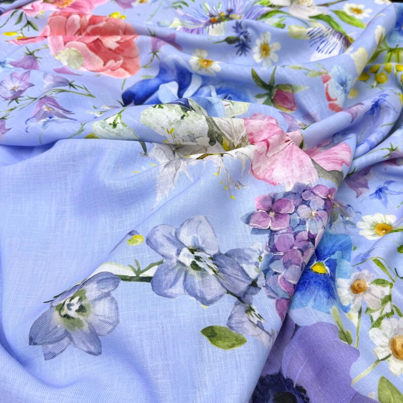 Peut inclure: Un tissu bleu clair orn&eacute; d'un motif floral. Le design pr&eacute;sente diverses fleurs aux tons rose, violet, bleu et blanc. Le tissu est drap&eacute;, mettant en valeur l'impression d&eacute;taill&eacute;e et la texture.