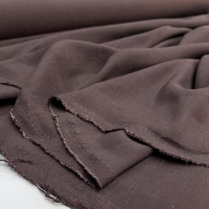 Peut inclure: Gros plan sur un tissu en lin marron foncé avec un bord brut et non fini. Le tissu est drapé en plis souples, révélant la texture et le tissage du matériau. La couleur est une teinte riche et terreuse, adaptée à divers projets de couture.