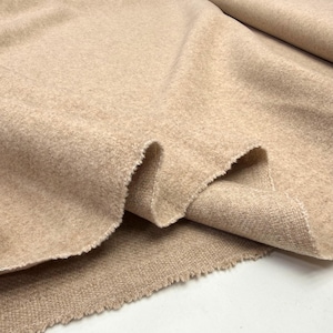 Puede incluir: Primer plano de una tela de color beige con una superficie texturizada y ligeramente difusa. La tela está doblada, revelando un reverso tejido y un borde en bruto sin terminar. El material parece suave y cálido, adecuado para ropa o decoración del hogar.