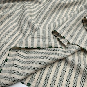 May include: Cream-colored fabric with dark green stripes. The stripes run diagonally across the material, creating a textured pattern. The edges of the fabric are visible, with a green trim. The fabric appears to be folded over itself.