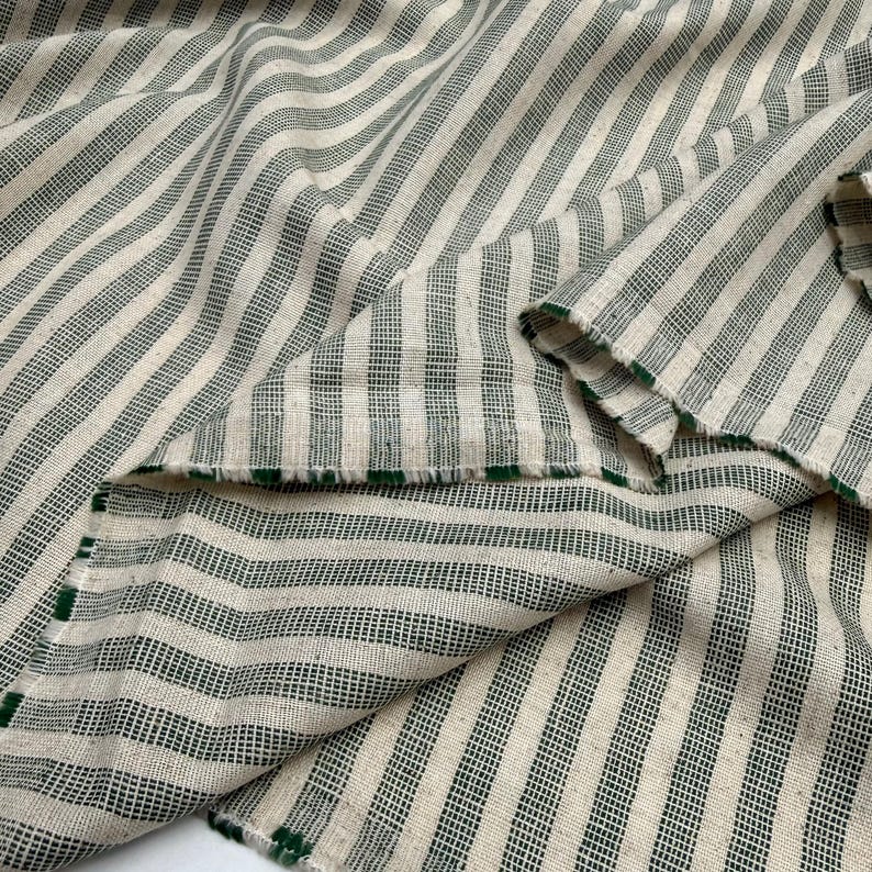May include: Cream-colored fabric with dark green stripes. The stripes run diagonally across the material, creating a textured pattern. The edges of the fabric are visible, with a green trim. The fabric appears to be folded over itself.