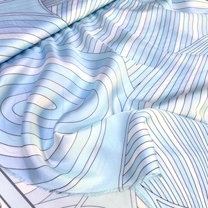 Op de afbeelding: Een stuk stof met een lichtblauwe en witte gradiëntachtergrond met een herhalend patroon van dunne, zwarte, gebogen en rechte lijnen. Het materiaal heeft een glad, licht reflecterend oppervlak met zichtbare plooien en een rafelige rand.