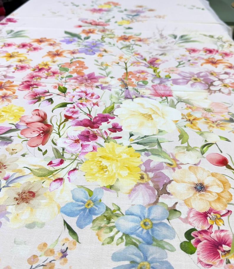 May include: A floral pattern tablecloth featuring a variety of colorful flowers in shades of pink, yellow, orange, purple, and blue. The flowers are arranged on a white background, creating a vibrant and cheerful design. The tablecloth is likely made of a soft fabric.