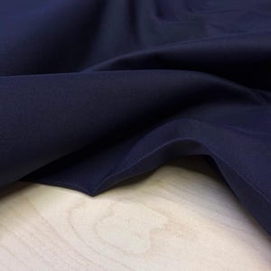May include: Close-up of navy blue fabric with visible texture and folds. The fabric is draped over a light-colored wooden surface, creating shadows and highlighting the material's texture. The deep blue color is consistent throughout.