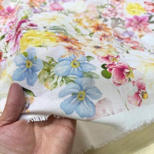 May include: White fabric with a vibrant floral pattern. The design features a variety of flowers in shades of pink, yellow, orange, and blue. The fabric is held in a hand, showing the raw edge of the material.