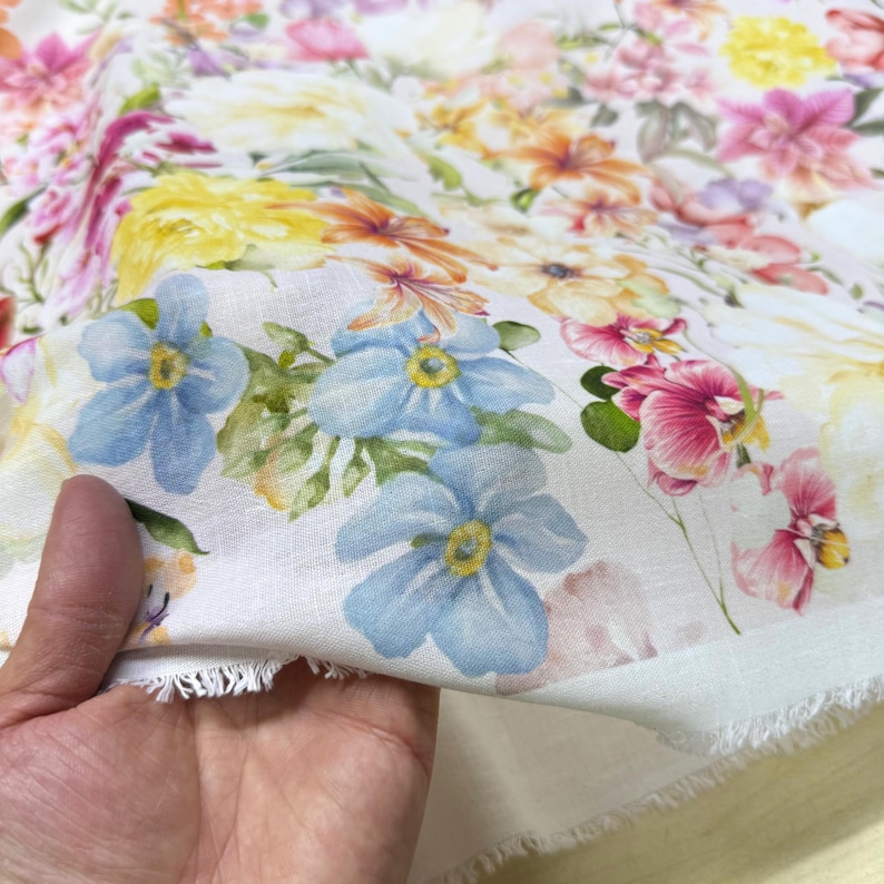 May include: White fabric with a vibrant floral pattern. The design features a variety of flowers in shades of pink, yellow, orange, and blue. The fabric is held in a hand, showing the raw edge of the material.