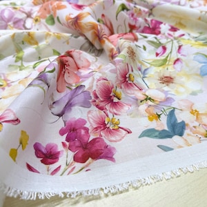 May include: Close-up of a white fabric with a vibrant floral pattern. The design features a variety of flowers in shades of pink, purple, yellow, and orange. The fabric has a raw edge, suggesting it's a cut piece, possibly for sewing or crafting.