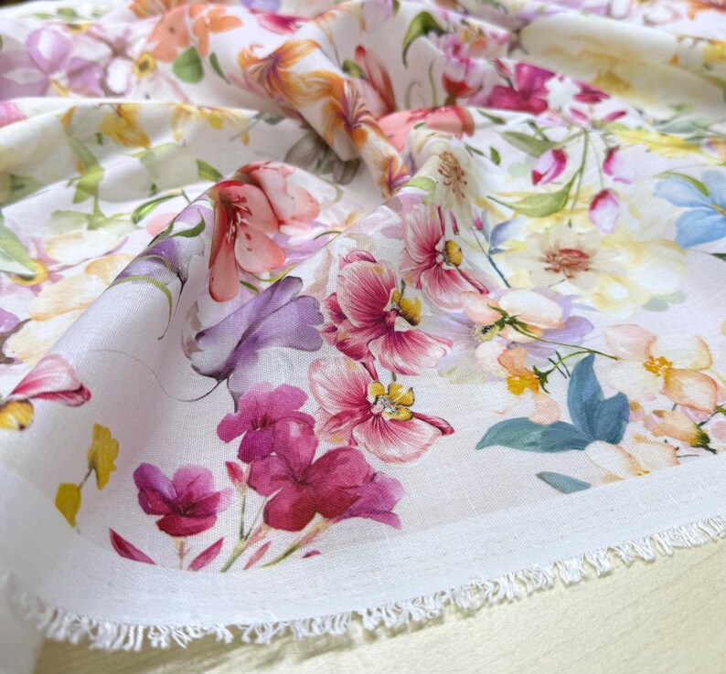 May include: Close-up of a white fabric with a vibrant floral pattern. The design features a variety of flowers in shades of pink, purple, yellow, and orange. The fabric has a raw edge, suggesting it's a cut piece, possibly for sewing or crafting.
