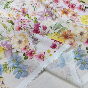 May include: White floral fabric featuring a watercolor design of various flowers in shades of pink, yellow, orange, and blue. The fabric has a raw edge and a soft, delicate appearance, suitable for crafting or home decor projects.