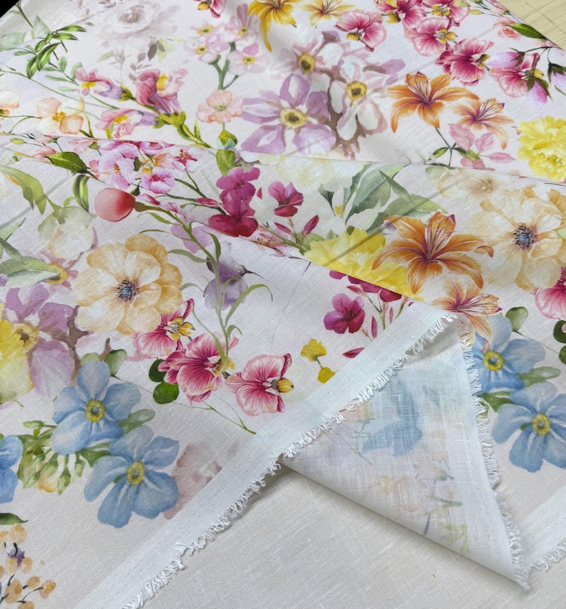 May include: White floral fabric featuring a watercolor design of various flowers in shades of pink, yellow, orange, and blue. The fabric has a raw edge and a soft, delicate appearance, suitable for crafting or home decor projects.