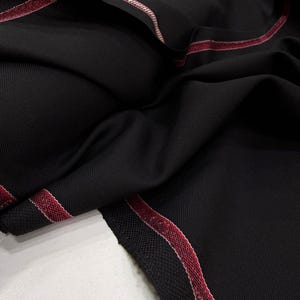 Może przedstawiać: Zbliżenie czarnej tkaniny o subtelnej prążkowanej fakturze. Krawędzie wykończone kontrastującym czerwono-białym paskiem. Tkanina jest udrapowana, ukazując fałdy i cienie.