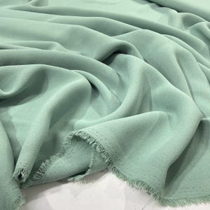 May include: A close-up shot of a soft, flowing fabric in a muted, mint green color. The fabric is draped and folded, with a textured surface and a frayed edge. The material appears lightweight and suitable for apparel or home decor.