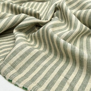 May include: Close-up of a folded fabric with a cream-colored background and vertical stripes in a muted green hue. The fabric has a textured appearance and a raw edge, suggesting it's a textile for sewing or crafting projects.
