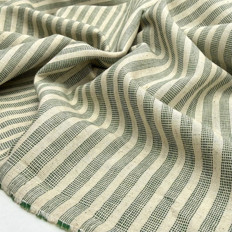 May include: Close-up of a folded fabric with a cream-colored background and vertical stripes in a muted green hue. The fabric has a textured appearance and a raw edge, suggesting it's a textile for sewing or crafting projects.