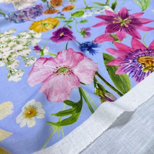 Peut inclure: Un tissu floral bleu clair avec une vari&eacute;t&eacute; de fleurs color&eacute;es. Le motif comprend des coquelicots roses, des passiflores violettes, des fleurs jaunes et des marguerites blanches. Le tissu a un bord de lisi&egrave;re blanc.