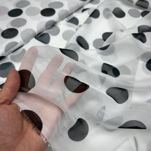 Puede incluir: Una tela blanca transparente con un patrón repetitivo de puntos irregulares y redondeados en gris oscuro y negro. El material es translúcido, permitiendo que la luz pase a través del tejido fino y ligero con una textura suave y drapeada.