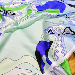 Pode incluir: Close-up de um tecido com um design floral abstrato colorido. O tecido apresenta um fundo azul claro com padrões em espiral em verde, azul, preto e branco. O tecido tem uma aparência macia e fluida.