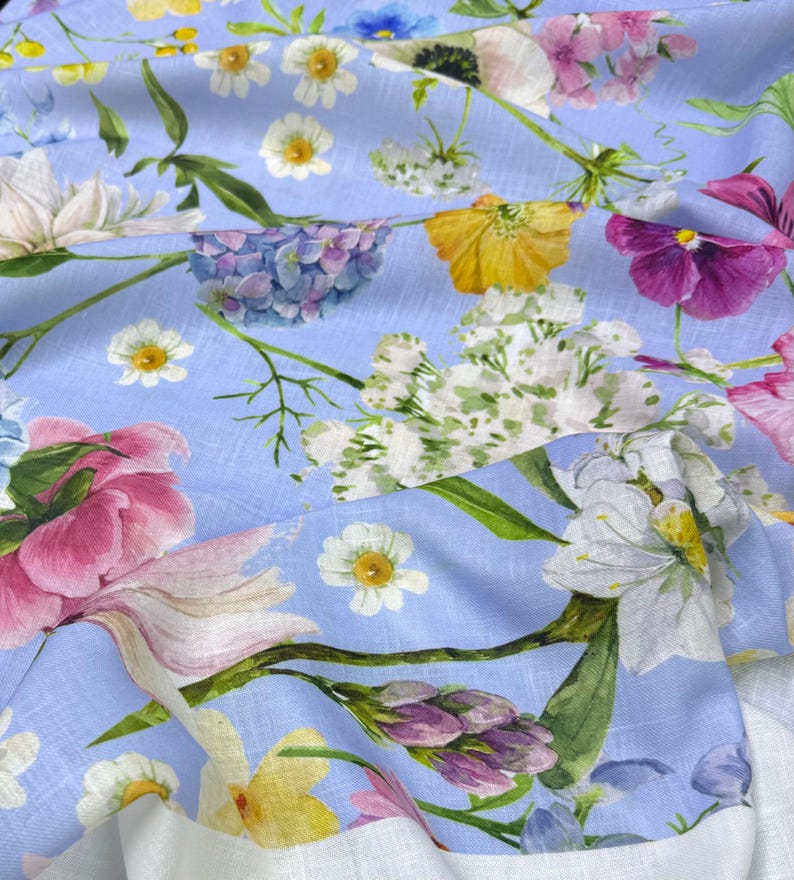 Peut inclure: Gros plan sur un tissu bleu clair orn&eacute; de motifs floraux aux couleurs rose, jaune, blanc et violet. Le tissu pr&eacute;sente une texture de type lin et une bordure blanche.