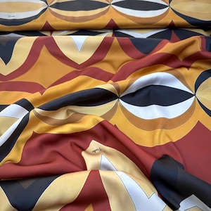 May include: A fabric featuring a repeating geometric pattern in shades of mustard yellow, deep red, black, and white. The design consists of interlocking curved shapes and pointed forms. The word Fendi is printed in small black text on the fabric.