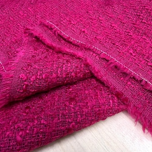 May include: Close-up of a vibrant fuchsia fabric with a textured weave. The fabric is folded, revealing the raw, fringed edges and a white stitch line. The material has a slightly irregular, nubby surface.