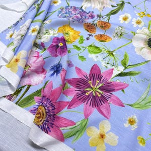 Peut inclure: Un tissu bleu clair orn&eacute; d'un motif floral vibrant. Le design pr&eacute;sente une vari&eacute;t&eacute; de fleurs aux tons roses, violets, jaunes et blancs, avec des feuilles vertes. Le tissu est pli&eacute;, r&eacute;v&eacute;lant un bord blanc.