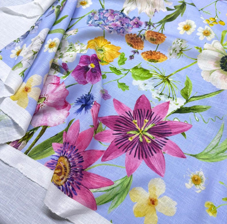 Peut inclure: Un tissu bleu clair orn&eacute; d'un motif floral vibrant. Le design pr&eacute;sente une vari&eacute;t&eacute; de fleurs aux tons roses, violets, jaunes et blancs, avec des feuilles vertes. Le tissu est pli&eacute;, r&eacute;v&eacute;lant un bord blanc.