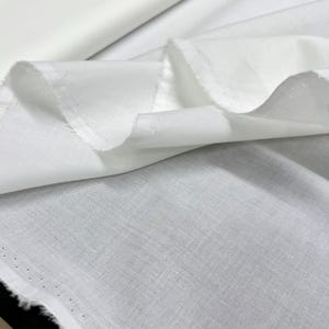 Könnte beinhalten: Nahaufnahme eines weißen Stoffes mit strukturiertem Gewebe. Der Stoff ist gefaltet und drapiert, wodurch die Kanten und die Textur sichtbar werden. Der Stoff scheint leicht und für Kleidung oder Heimdekoration geeignet zu sein.
