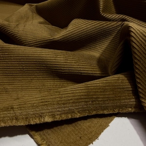 Puede incluir: Primer plano de una tela de pana verde oliva doblada. La tela tiene una textura acanalada y un aspecto suave y aterciopelado. Los bordes están deshilachados, mostrando la textura del material. Adecuado para ropa y tapicería.