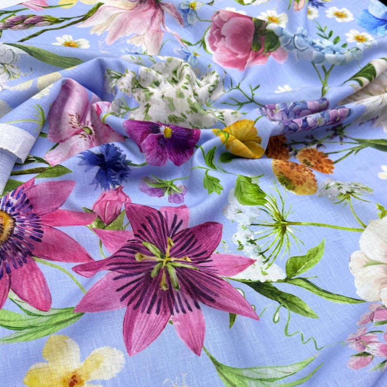 Peut inclure: Un tissu bleu clair orn&eacute; d'un motif floral vibrant. Le design pr&eacute;sente une vari&eacute;t&eacute; de fleurs dans les tons de rose, violet, jaune et blanc, avec des feuilles et des tiges vertes. Le tissu est probablement en coton ou en lin.