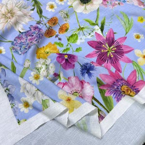 Peut inclure: Un tissu bleu clair orn&eacute; d'un motif floral color&eacute; avec des fleurs roses, violettes, jaunes et blanches. Le tissu a une bordure blanche et est pli&eacute;, r&eacute;v&eacute;lant le motif floral d&eacute;taill&eacute; de style aquarelle. Le tissu est probablement en lin ou dans une mati&egrave;re similaire.