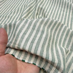 May include: Close-up of a green and white striped fabric. The fabric has vertical stripes of varying widths. The material appears to be a woven cotton or linen, with a slightly textured surface. The edges of the fabric are frayed.