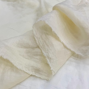 Puede incluir: Una pieza de tela color blanco roto con un tejido texturizado visible y bordes deshilachados. El material está drapeado en pliegues suaves, resaltando la calidad semitransparente y las sutiles líneas horizontales irregulares del tejido.
