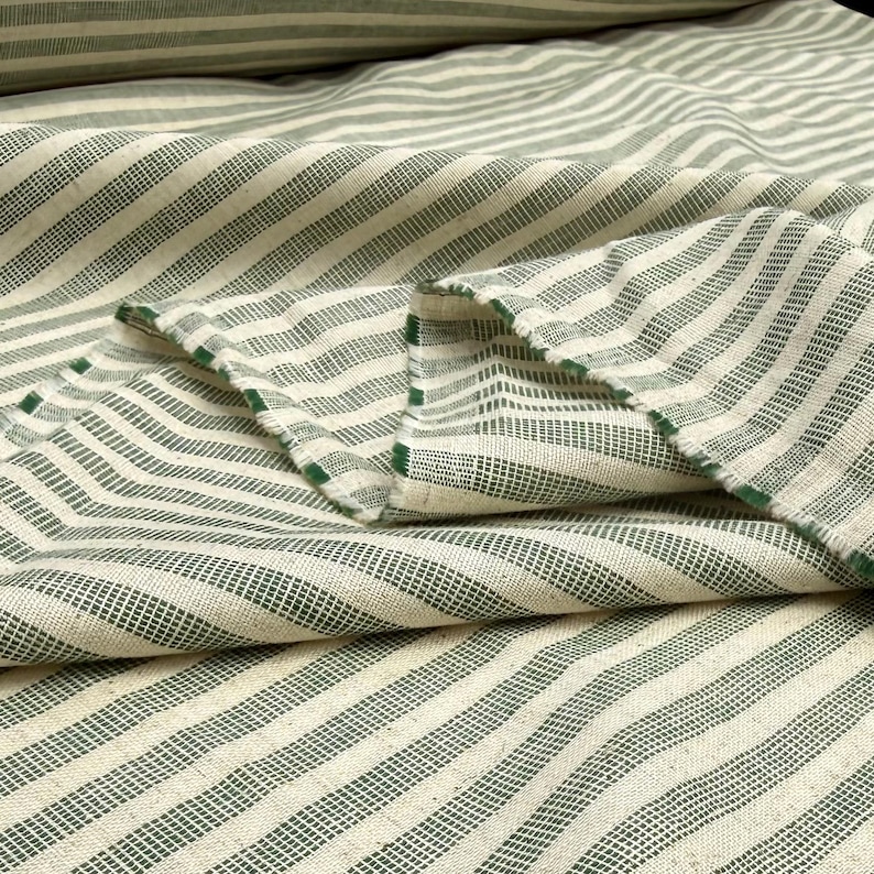 May include: Close-up of a folded fabric with a cream-colored background and green stripes. The stripes run diagonally across the fabric. The edges of the fabric are frayed, revealing the green thread. The fabric appears to be a woven material.