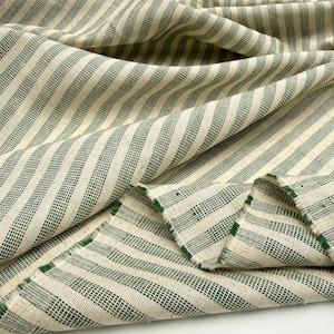 May include: Close-up of a cream-colored fabric with a green striped pattern. The fabric is folded and draped, showing the texture and weave of the material. The stripes are evenly spaced and run diagonally across the fabric.