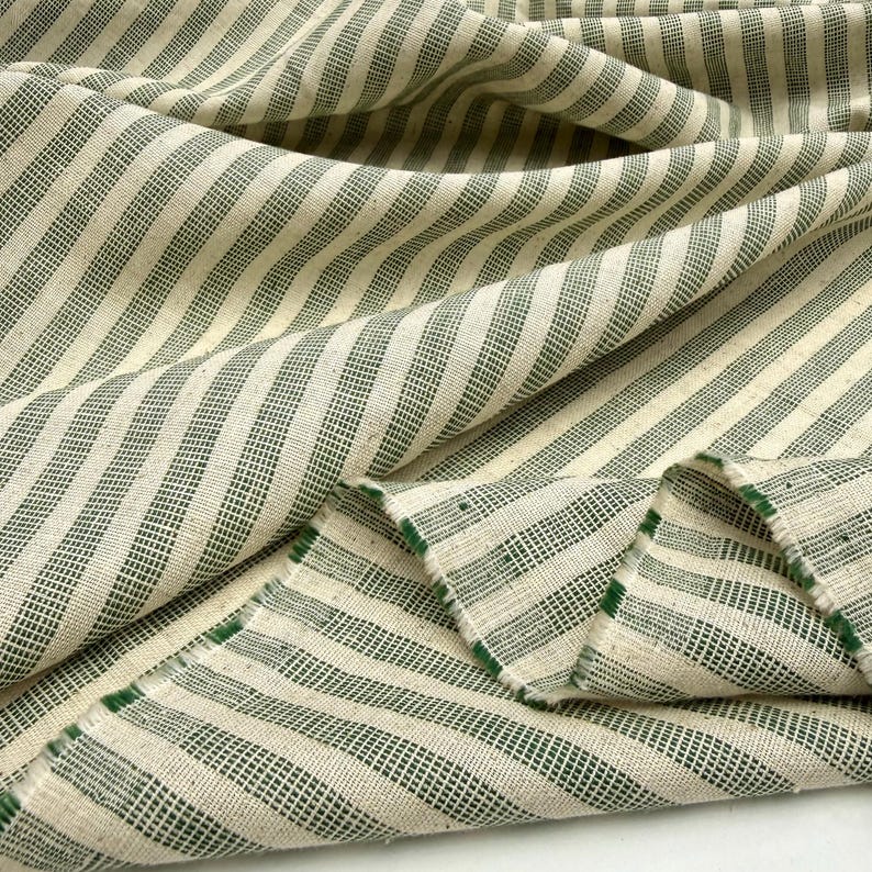 May include: Close-up of a cream-colored fabric with a green striped pattern. The fabric is folded and draped, showing the texture and weave of the material. The stripes are evenly spaced and run diagonally across the fabric.