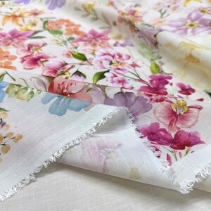 May include: Close-up of a white fabric with a vibrant floral pattern. The design features watercolor-style flowers in shades of pink, purple, orange, and blue, with green leaves. The fabric has a raw, fringed edge, suggesting it's a cut piece.