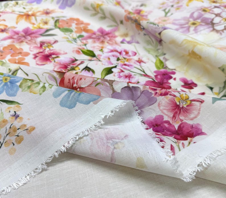 May include: Close-up of a white fabric with a vibrant floral pattern. The design features watercolor-style flowers in shades of pink, purple, orange, and blue, with green leaves. The fabric has a raw, fringed edge, suggesting it's a cut piece.