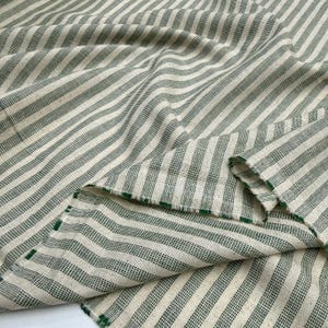 May include: Cream-colored fabric with a green striped pattern. The stripes are evenly spaced and run diagonally across the fabric. The fabric is folded over in places, showing the texture and the green edge.