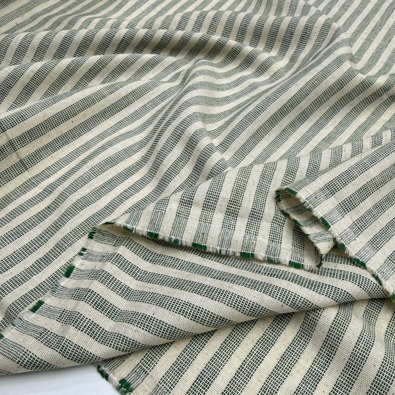 May include: Cream-colored fabric with a green striped pattern. The stripes are evenly spaced and run diagonally across the fabric. The fabric is folded over in places, showing the texture and the green edge.