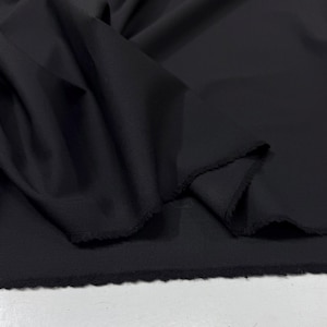 Op de afbeelding: Close-up van zwarte stof. De stof is gedrapeerd en gevouwen, waardoor de textuur en de onafgewerkte randen zichtbaar zijn. Het materiaal lijkt zacht en soepel.