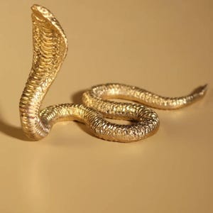 Könnte beinhalten: Eine goldfarbene Kobraskulptur mit strukturierter Oberfläche. Die Schlange ist in einer auffälligen, aufrechten Position dargestellt, mit aufgestelltem Kopf. Die Skulptur befindet sich vor einem einfarbigen, neutralen Hintergrund.