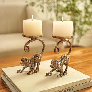 May include: Two brass-colored monkey candle holders with white candles. The monkey figures are detailed and posed as if crawling. The candle holders are on a stack of books on a wooden table.
