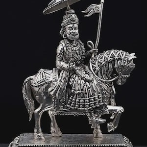 May include: A detailed silver statue of a figure riding a horse, holding a flag. The figure wears ornate clothing and a headdress, and is sheltered by a parasol. The statue sits on a rectangular base with decorative edges. The words "exotic india" are visible.
