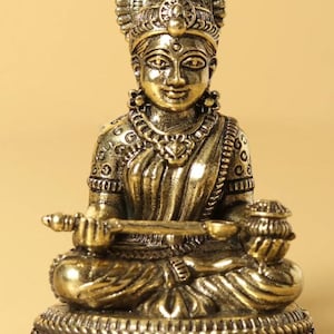 May include: A gold-colored statue of a seated deity, adorned with intricate details. The figure wears a crown and jewelry, holding a musical instrument and a small pot. The statue is on a decorative base, with a serene expression.