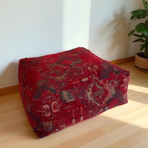 Könnte beinhalten: Ein quadratischer, roter Pouf mit einem detaillierten, gemusterten Design in Braun-, Schwarz- und Beigetönen. Der Pouf besteht aus einem strukturierten Stoff und sieht aus wie ein Bodenkissen oder eine Ottomane. Das Design ähnelt einem traditionellen Teppich.