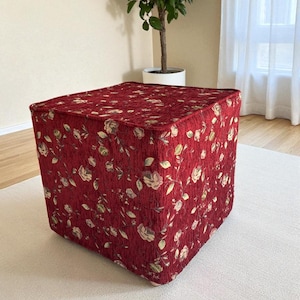 Puede incluir: Un otomán cuadrado con un estampado floral rojo. La tela presenta un diseño repetido de flores y hojas crema y verdes sobre un fondo rojo oscuro. El otomán tiene forma de cubo.